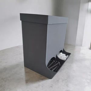 May include: A matte dark gray storage bin featuring two circular front openings at the base. The left opening contains several black metal bolts, while the right opening holds a collection of small white plastic fasteners or wall anchors.