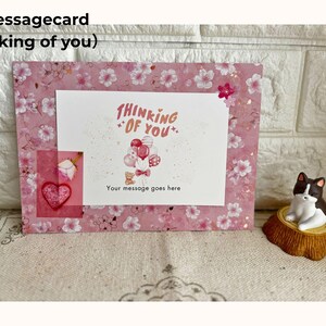 May include: A pink message card with a floral pattern and the words "Thinking of You." The card features a white insert with a balloon and teddy bear illustration. A small heart and rose are visible.