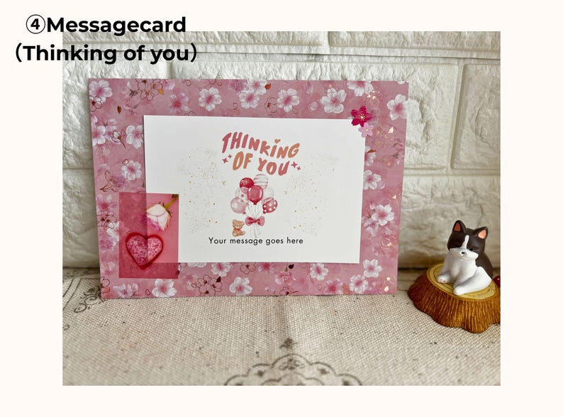 May include: A pink message card with a floral pattern and the words "Thinking of You." The card features a white insert with a balloon and teddy bear illustration. A small heart and rose are visible.