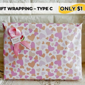 May include: Gift-wrapped package with a heart pattern in shades of pink and peach. A pink ribbon and a small tag with a floral design adorn the package. The text "GIFT WRAPPING - TYPE C" is at the top.