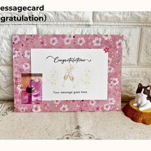 May include: A pink message card with a floral pattern and the word "Congratulations" in script. The card features an illustration of champagne glasses and the text "Your message goes here". A small cat figurine sits nearby.