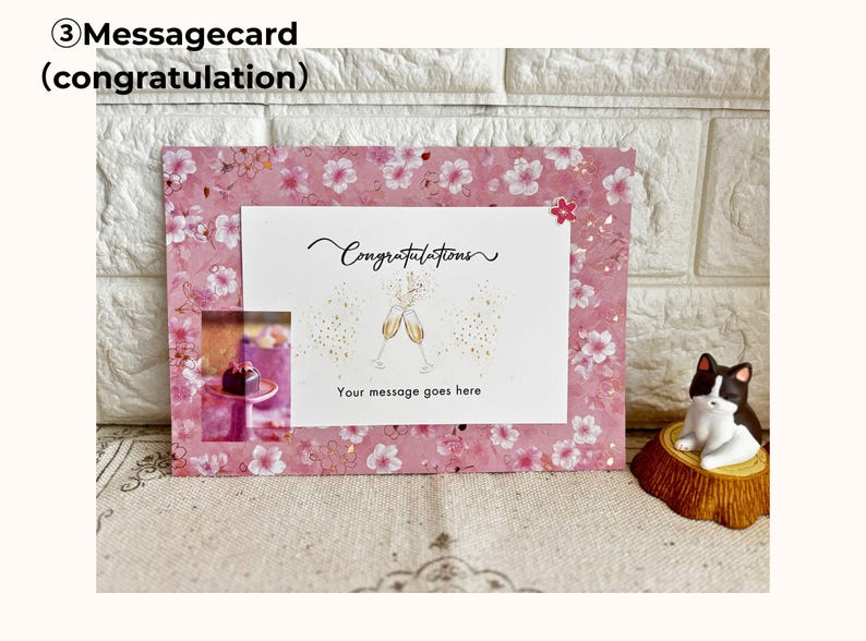 May include: A pink message card with a floral pattern and the word "Congratulations" in script. The card features an illustration of champagne glasses and the text "Your message goes here". A small cat figurine sits nearby.