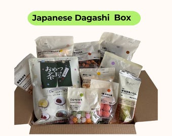 May include: A cardboard box filled with various Japanese snacks, including packaged candies, cookies, and other treats. The box is labeled "Japanese Dagashi Box" in a green banner above. The snacks are in white and clear packaging.