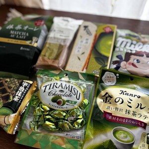May include: Assortment of Japanese matcha-flavored snacks, including matcha au lait, tiramisu chocolate, and matcha latte candies. The packaging features green and gold colors, with Japanese text and illustrations. A variety of treats are displayed on a light-colored surface.
