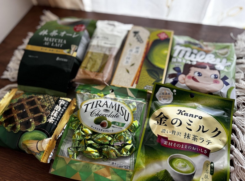 May include: Assortment of Japanese matcha-flavored snacks, including matcha au lait, tiramisu chocolate, and matcha latte candies. The packaging features green and gold colors, with Japanese text and illustrations. A variety of treats are displayed on a light-colored surface.
