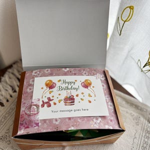 May include: A gift box with a floral patterned lid and a white card that reads "Happy Birthday!" with a cake and balloons. The box is open, revealing the contents inside. The box is sitting on a patterned surface.