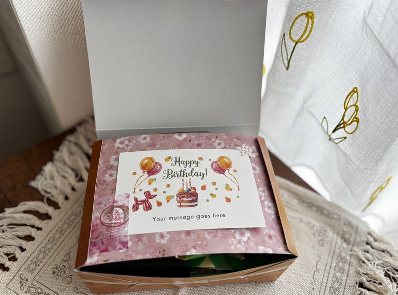 May include: A gift box with a floral patterned lid and a white card that reads "Happy Birthday!" with a cake and balloons. The box is open, revealing the contents inside. The box is sitting on a patterned surface.