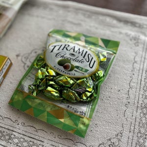 May include: A package of Tiramisu Chocolate candies in green foil wrappers. The package has a white oval label with the words "TIRAMISU Chocolate" and a graphic of a green nut. Other packaging is visible in the background.