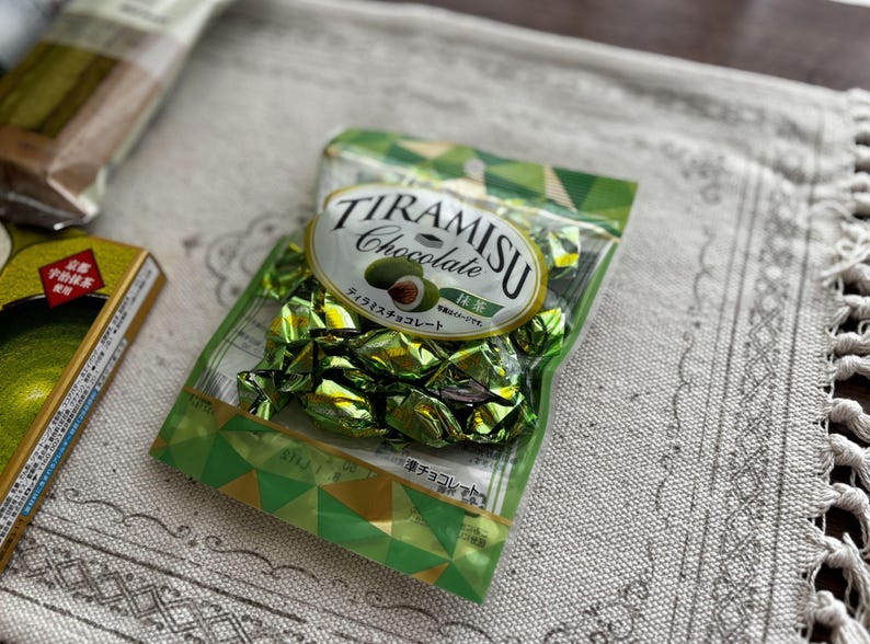 May include: A package of Tiramisu Chocolate candies in green foil wrappers. The package has a white oval label with the words "TIRAMISU Chocolate" and a graphic of a green nut. Other packaging is visible in the background.