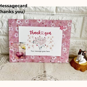 May include: A pink floral message card with the words "thank you" and a bouquet illustration. The card has a small window with a heart-shaped object. A small cat figurine sits to the right.