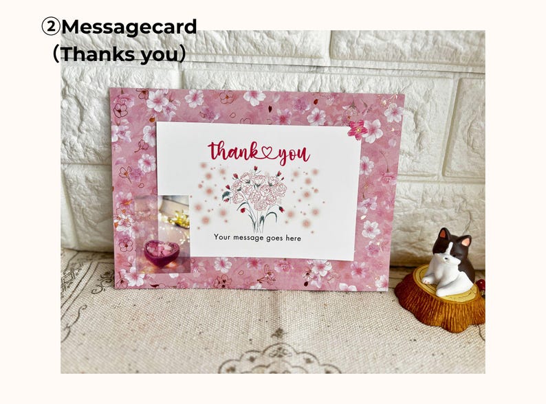 May include: A pink floral message card with the words "thank you" and a bouquet illustration. The card has a small window with a heart-shaped object. A small cat figurine sits to the right.
