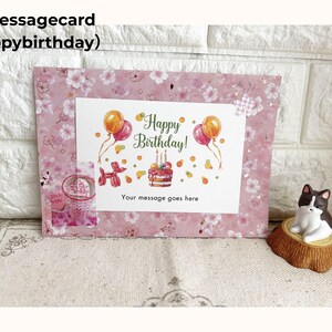 May include: A pink message card with a floral pattern and the text "Happy Birthday!" featuring balloons and a cake illustration. The card includes space for a personalized message. A small cat figurine sits nearby.