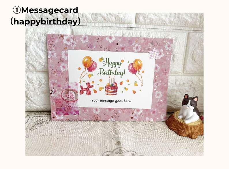 May include: A pink message card with a floral pattern and the text "Happy Birthday!" featuring balloons and a cake illustration. The card includes space for a personalized message. A small cat figurine sits nearby.