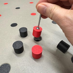 以下が含まれることがあります： 赤と黒のペグが付いたペグボードのクローズアップ。手は赤いペグの上にネジを持っています。