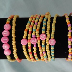 Könnte beinhalten: Armbänder aus gelben, rosa, orangefarbenen und türkisfarbenen Perlen. Ein Armband schreibt "BRIDE" mit rosa Buchstabenperlen. Andere Armbänder schreiben "TEAM" mit rosa Buchstabenperlen. Die Armbänder werden auf einer schwarzen Oberfläche präsentiert.