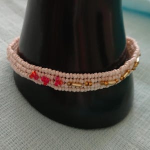 Könnte beinhalten: Ein handgefertigtes Armband mit einem gewebten, cremefarbenen Band. Das Armband ist mit kleinen, roten Blumenmotiven und goldfarbenen Perlen verziert. Es wird auf einem schwarzen, zylindrischen Ständer präsentiert, der sein aufwendiges Design hervorhebt.