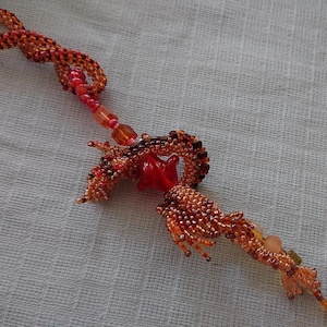 May include: A beaded dragon pendant in shades of orange, red, and brown. The dragon features a detailed design with a red glass centerpiece. The pendant has a loop at the top and a beaded tail, perfect for a necklace or keychain.