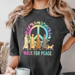 以下が含まれることがあります： 虹色のグラデーションの平和のシンボルが描かれた濃い灰色のTシャツ。周りには花が描かれています。シンボルの下には「WALK FOR PEACE」の文字。ローブを着た人物と犬がデザインされています。