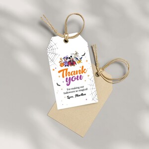 May include: A white rectangular gift tag with a brown twine loop, featuring a Halloween-themed illustration of cartoon characters in costumes, spider webs, and bats. The text reads: Thank you, For making our halloween so magical!, Love, Martlen.