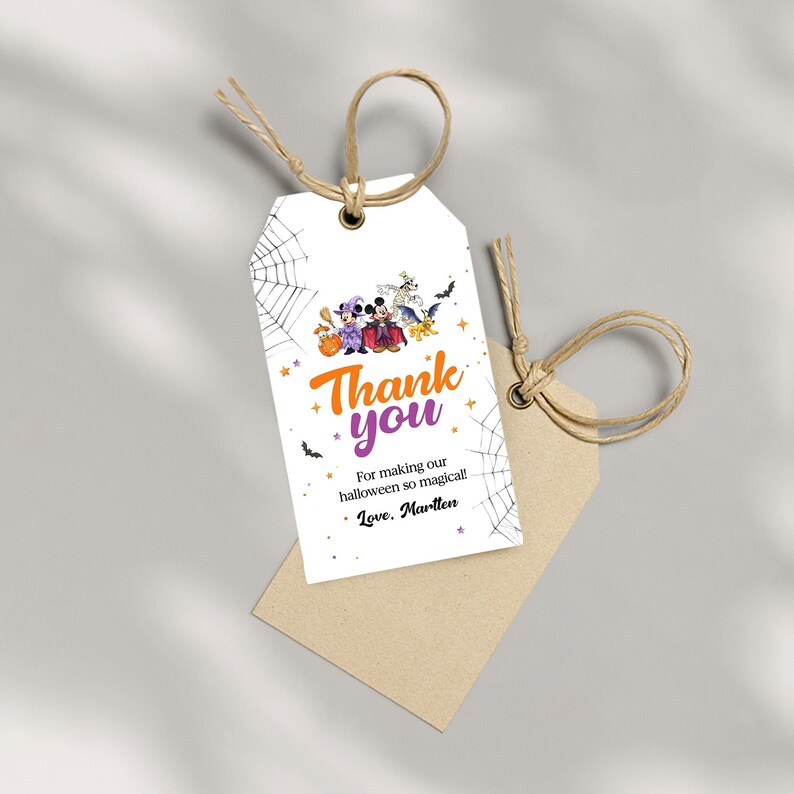 May include: A white rectangular gift tag with a brown twine loop, featuring a Halloween-themed illustration of cartoon characters in costumes, spider webs, and bats. The text reads: Thank you, For making our halloween so magical!, Love, Martlen.