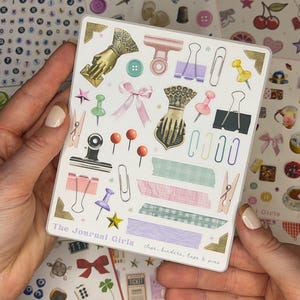 May include: A sheet of stickers featuring various office supplies, including clips, binders, tape, and pins. The stickers are illustrated in a vintage style with pastel colors and gold accents. The sheet is labeled "The Journal Girls" and includes the text "clips, binders, tape & pins".