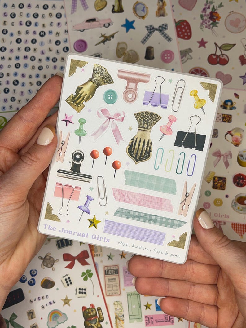 May include: A sheet of stickers featuring various office supplies, including clips, binders, tape, and pins. The stickers are illustrated in a vintage style with pastel colors and gold accents. The sheet is labeled "The Journal Girls" and includes the text "clips, binders, tape & pins".