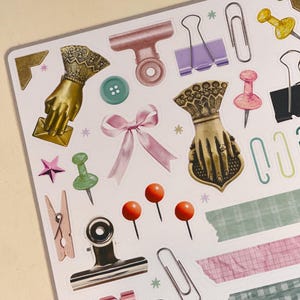 May include: A sheet of stickers featuring various office supplies, including paper clips, binder clips, push pins, and decorative elements. The stickers are on a white background.