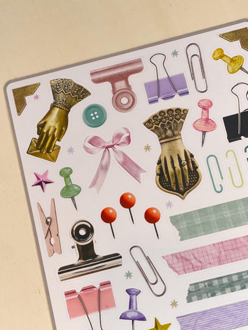 May include: A sheet of stickers featuring various office supplies, including paper clips, binder clips, push pins, and decorative elements. The stickers are on a white background.
