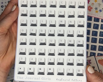 Könnte beinhalten: Ein Blatt mit weißen Aufklebern mit schwarzen Buchstaben und Symbolen, die die Tasten einer Tastatur darstellen. Die Aufkleber sind in Reihen und Spalten angeordnet, und der Text "The Journal Girls keyboard letters" ist unten auf dem Blatt gedruckt.