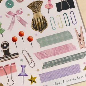 May include: A sheet of colorful stickers featuring various office supplies, including paper clips, binders, tape, and push pins. The stickers are designed in a vintage style with a light background and pastel colors. The text "The Journal Girls" and "clips, binders, tape &" are printed on the sheet.