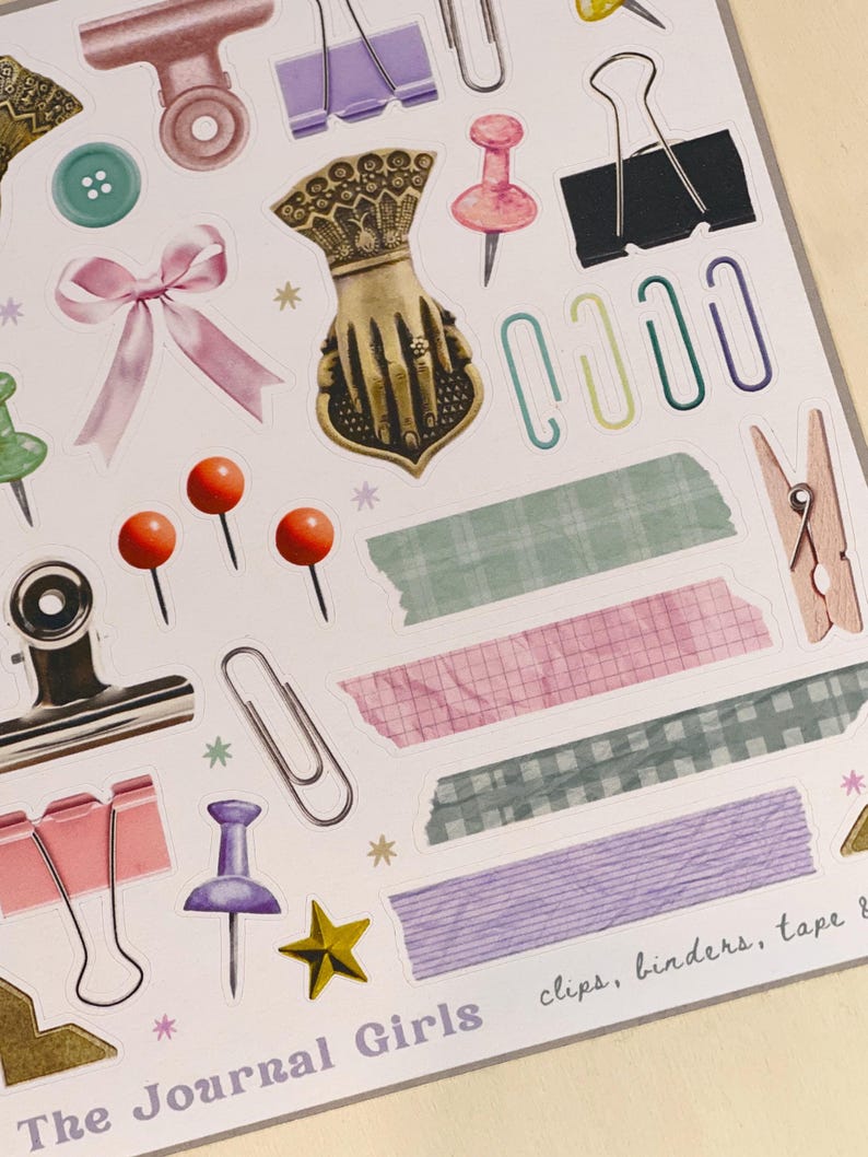 May include: A sheet of colorful stickers featuring various office supplies, including paper clips, binders, tape, and push pins. The stickers are designed in a vintage style with a light background and pastel colors. The text "The Journal Girls" and "clips, binders, tape &" are printed on the sheet.