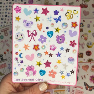 May include: A sheet of colorful stickers featuring a variety of designs, including stars, hearts, animals, and smiley faces. The stickers are on a white background with a pink border. The text "The Journal Girls girlhood stickers & sequins" is printed at the bottom of the sheet.