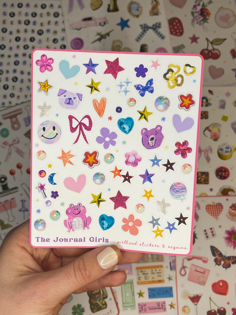 May include: A sheet of colorful stickers featuring a variety of designs, including stars, hearts, animals, and smiley faces. The stickers are on a white background with a pink border. The text "The Journal Girls girlhood stickers & sequins" is printed at the bottom of the sheet.