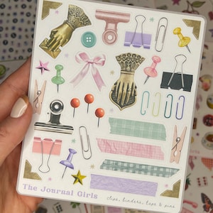 May include: A sheet of stickers featuring various office supplies, including clips, binders, tape, and pins. The stickers are illustrated in a vintage style with pastel colors and gold accents. The text "The Journal Girls" and "clips, binders, tape & pins" are printed on the sheet.