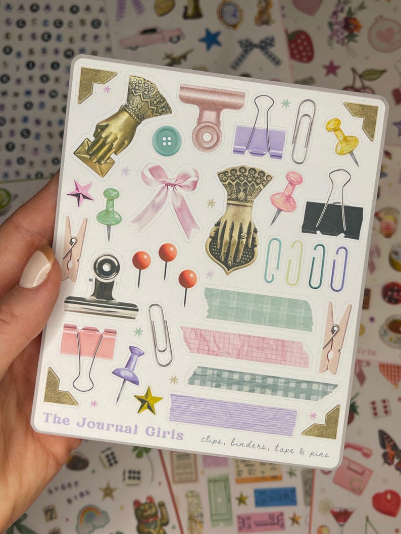 May include: A sheet of stickers featuring various office supplies, including clips, binders, tape, and pins. The stickers are illustrated in a vintage style with pastel colors and gold accents. The text "The Journal Girls" and "clips, binders, tape & pins" are printed on the sheet.