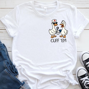 May include: White t-shirt featuring a cartoon graphic of two ducks. One duck is wearing a nurse's hat and sunglasses, holding a blood pressure cuff. The other duck looks surprised. The text "CUFF 'EM" is below the ducks.