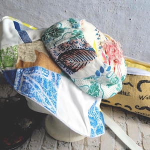 Peut inclure: Casquette de style gavroche en patchwork avec divers motifs de tissus, dont des motifs floraux, géométriques et des couleurs unies. La casquette a un bord blanc et une sangle. Un sac jaune avec le mot "ABLE" est visible en arrière-plan.