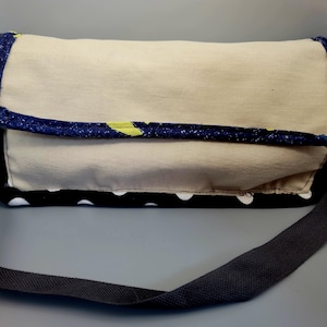 May include: A beige messenger bag with a flap closure. The bag has a navy blue trim with yellow accents and a black and white polka dot base. A dark grey strap is attached for carrying.