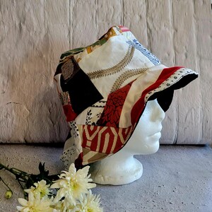 May include: A patchwork bucket hat constructed from various fabric scraps featuring a mix of white, red, black, and gold patterns. The hat has a wide, downward-sloping brim and is displayed on a white mannequin head with white flowers nearby.