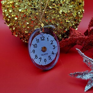 May include: An oval-shaped pendant necklace featuring a white clock face with black numerals from 1 to 12 and the word TALKING printed in the centre. The clock face is encased in a clear resin frame and hangs from a thin gold-toned metal chain.