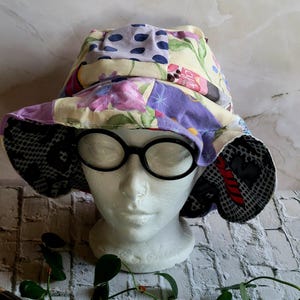 May include: A patchwork sun hat with a wide brim, featuring floral, polka dot, and geometric patterns in shades of yellow, purple, and black. The hat is displayed on a white mannequin head with black-rimmed glasses.