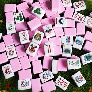 May include: A collection of rectangular mahjong tiles with pink backs and white fronts featuring various colorful illustrations, including soccer-themed graphics, maple leaves, cacti, eagles, and the word JOKER printed on several tiles.