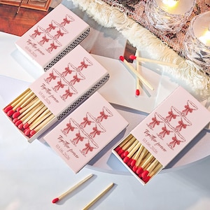 Może przedstawiać: Różowe pudełka z zapałkami z czerwonymi końcówkami, z motywem kieliszków do szampana i napisem "Together forever 03.06.2026." Widoczne są również dwa podświetlane, przezroczyste, fasetowane świeczniki.