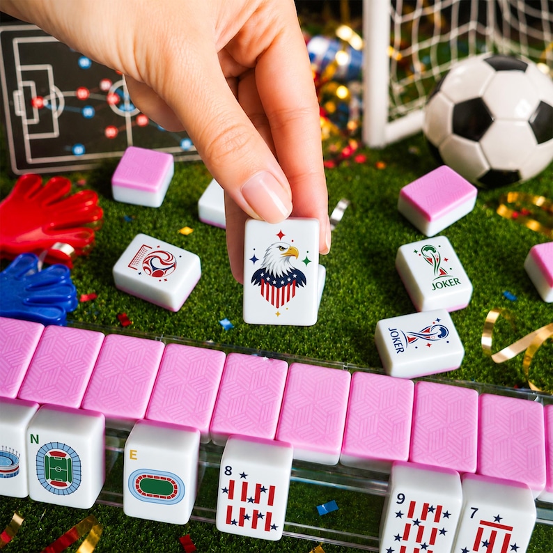 Puede incluir: Una ficha de mahjong blanca con un gr&aacute;fico de &aacute;guila calva con un patr&oacute;n de bandera roja, blanca y azul, sostenida sobre un conjunto de fichas rosas y blancas sobre una superficie verde. Otras fichas muestran gr&aacute;ficos de f&uacute;tbol y estadios.