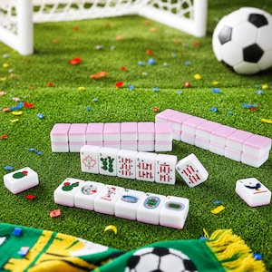 Puede incluir: Un conjunto de fichas de mahjong blancas y rosas con varios gr&aacute;ficos de tem&aacute;tica deportiva, incluyendo un bal&oacute;n de f&uacute;tbol, un estadio y una hoja de arce. Las fichas est&aacute;n dispuestas en filas y esparcidas sobre una superficie verde con confeti.