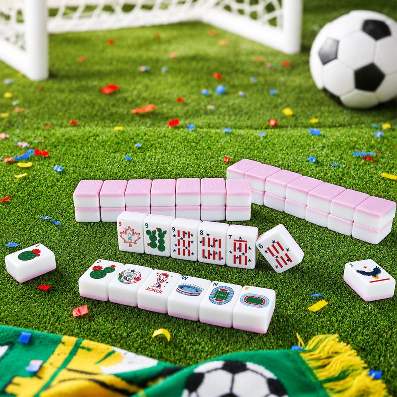 以下が含まれることがあります： サッカーボール、スタジアム、カエデの葉など、スポーツをテーマにした様々なグラフィックが描かれた白とピンクの麻雀牌セット。牌は列に並べられ、紙吹雪が散らばる緑色の表面の上に置かれており、近くにはサッカーボールがある。