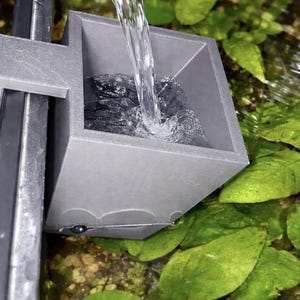 Puede incluir: Un filtro de acuario gris cuadrado con agua vertiéndose en él. El filtro está sujeto a un soporte negro y está rodeado de plantas acuáticas verdes. El agua es clara y fluye, creando un efecto visual dinámico.