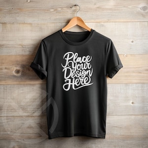 Könnte beinhalten: Schwarzes T-Shirt mit dem Text "Place Your Design Here" in weißer Schrift. Das Shirt hängt an einem Holzbügel vor einem Holzgrund. Die Ärmel sind umgeschlagen.