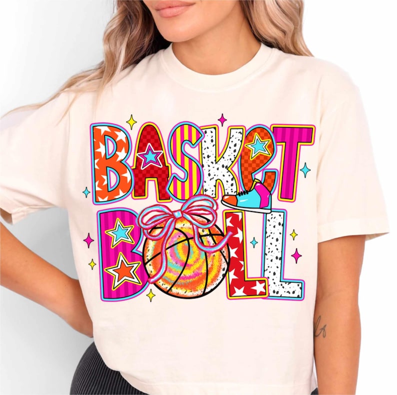 Op de afbeelding: Een wit T-shirt met een kleurrijk grafisch ontwerp met de tekst "BASKETBALL" met een basketbal, een schoen en sterren.