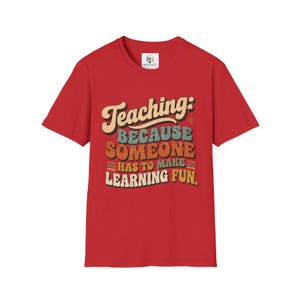 Op de afbeelding: Een rood t-shirt met korte mouwen en een meerkleurige grafische print met de tekst: Teaching: BECAUSE SOMEONE HAS TO MAKE LEARNING FUN. Het ontwerp gebruikt een retro-stijl lettertype met gelaagde kleuren, waaronder crème, blauwgroen en oranje.